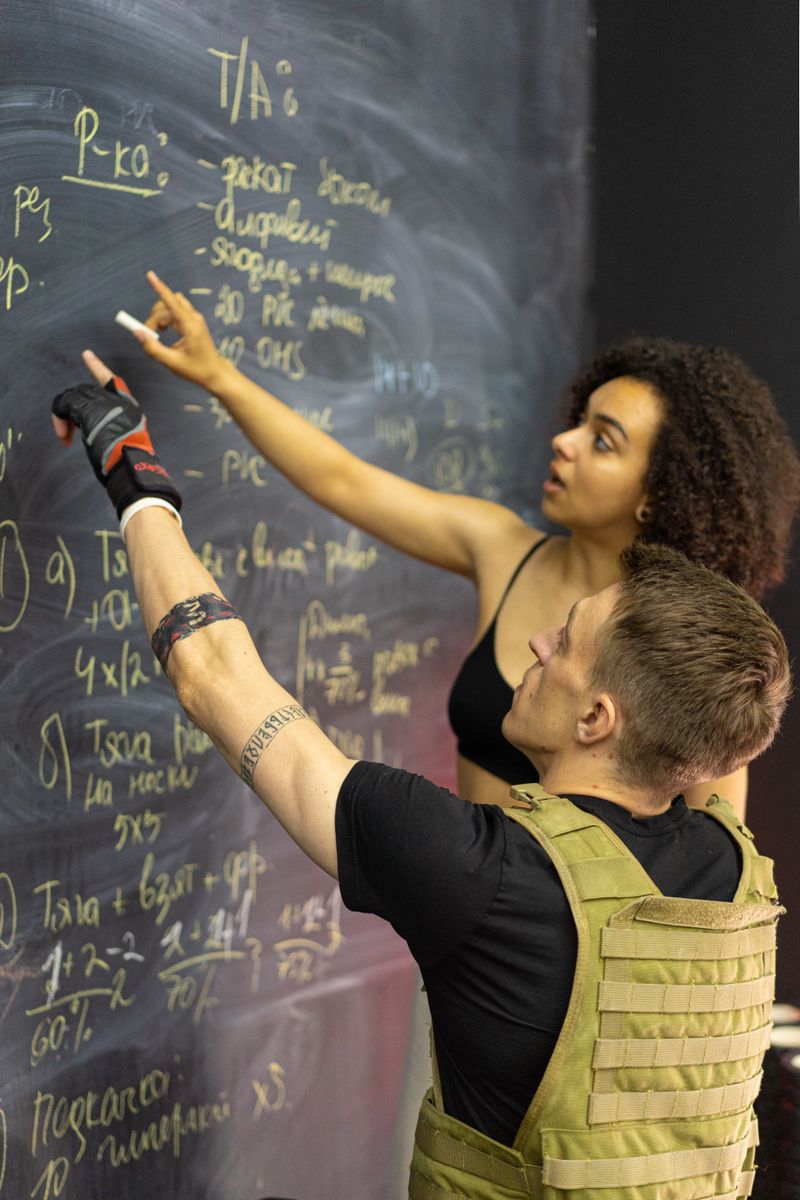 Two adults in fitness gear analyze workout plans on a chalkboard in a gym setting.