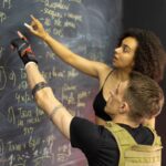 회원은 운동이 아니라 강사를 따라옵니다 | 필라테스,바레, 스텝박스자격증 Two adults in fitness gear analyze workout plans on a chalkboard in a gym setting.
