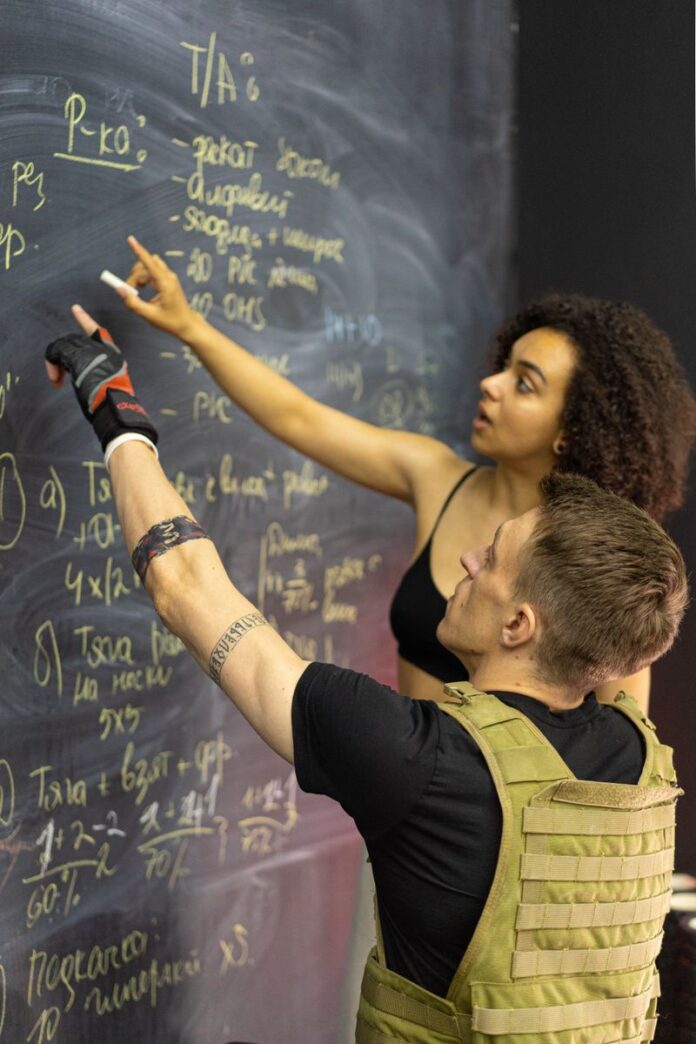 Photo by Mikhail Nilov Two adults in fitness gear analyze workout plans on a chalkboard in a gym setting.